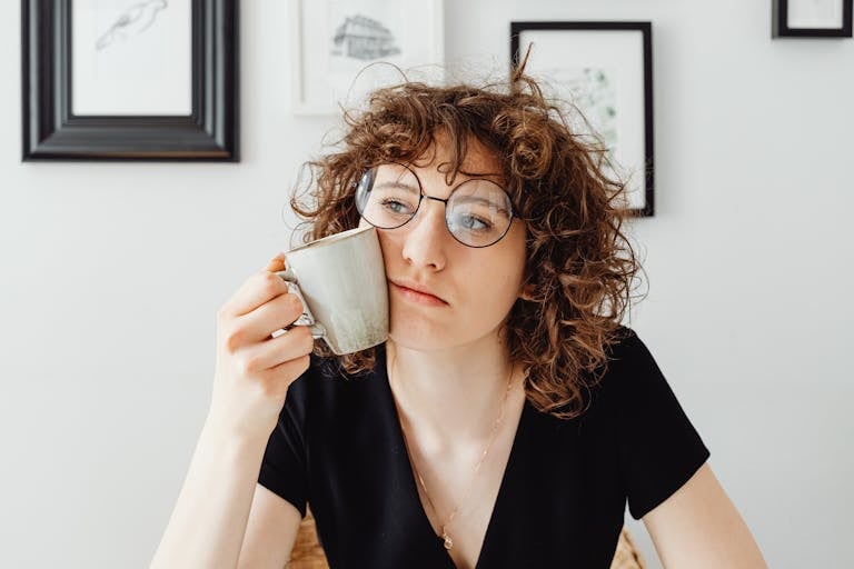 A thoughtful woman with curly hair and glasses holding a coffee mug in a modern room.