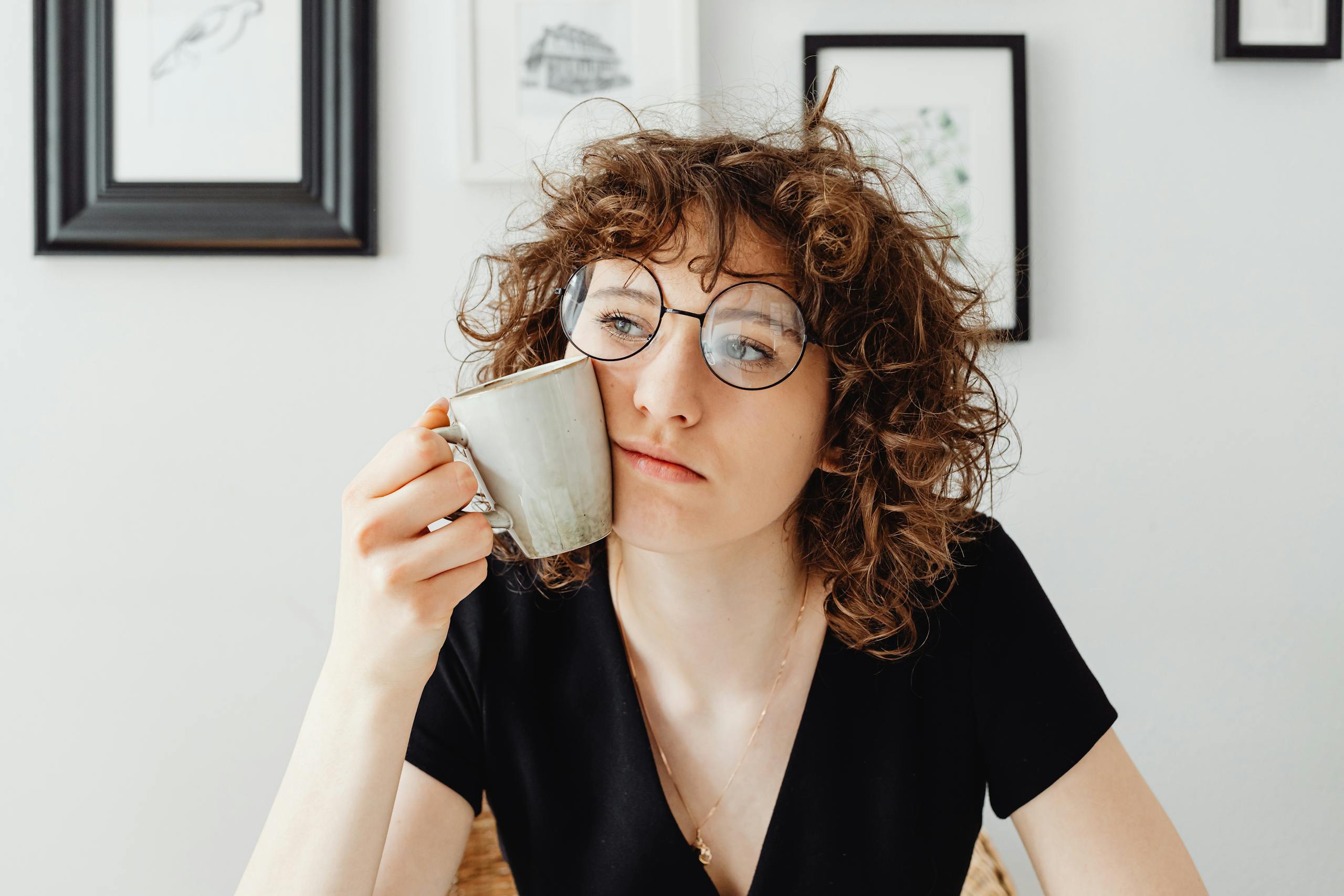 A thoughtful woman with curly hair and glasses holding a coffee mug in a modern room.